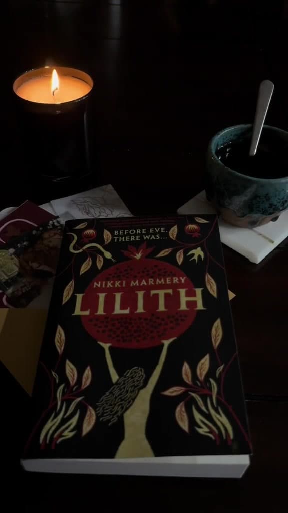 A copy of Lilith by Nikki Marmery on a dark table beside a lit candle and a cup of tea
